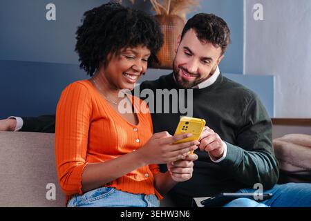 Happy diverse couple sitting on a sofa, looking at smartphone together. Horizontal. Stock Photo