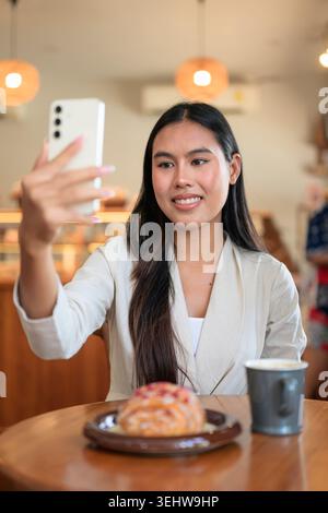 Vertical portrait of a confident young Isan Thai businesswoman smiling while taking a selfie or hosting a video call with her smartphone at a bakery c Stock Photo