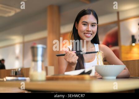 Horizontal eye-level medium portrait of a cheerful young Isan Thai woman smiling and making direct eye contact with the camera while lifting noodles w Stock Photo