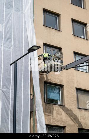 Construction worker on hydraulic lift platform repairing residential apartment building facade in urban area Stock Photo