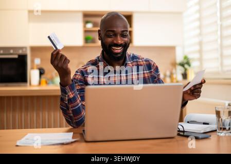 A cheerful man sits at his kitchen table with a laptop, credit card, and documents, suggesting successful online payment or financial management. Stock Photo