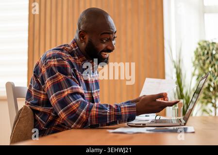 Happy black man is doing home budget accountancy. He is holding financial bills and using laptop. Stock Photo