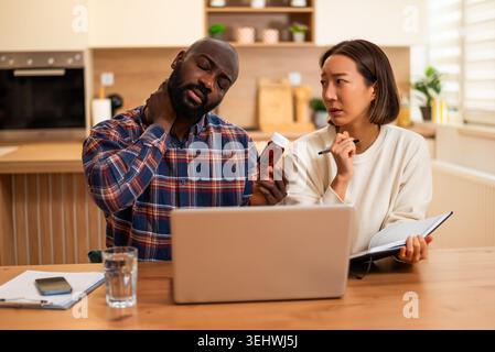 A couple sits at a table looking concerned while using a laptop, with the man holding his neck and the woman checking medication and taking notes. Stock Photo