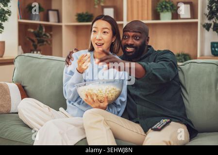 A joyful multiracial couple sits closely together on a green sofa, laughing and embracing while watching tv and eating popcorn. Stock Photo