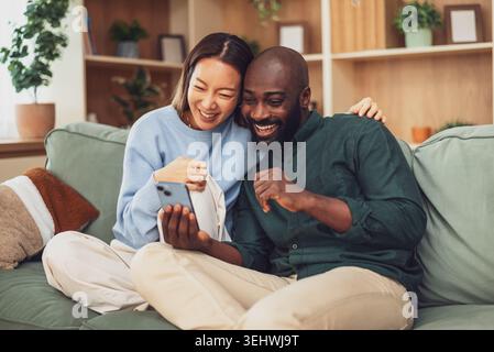 Happy and loving multiracial couple shares a joyful moment while relaxing at home. They are using smartphone. Stock Photo