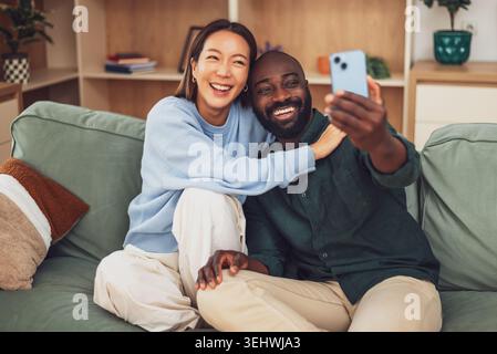A joyful multiracial couple sits closely together on a green sofa, laughing and embracing in a cozy, modern living room. They are taking selfie. Stock Photo