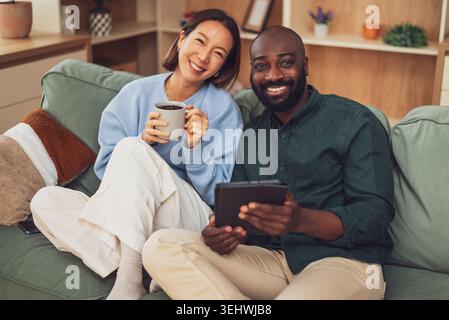 Happy interracial couple shares a joyful moment while relaxing at home. They are drinking coffee,talking and using digital tablet. Stock Photo