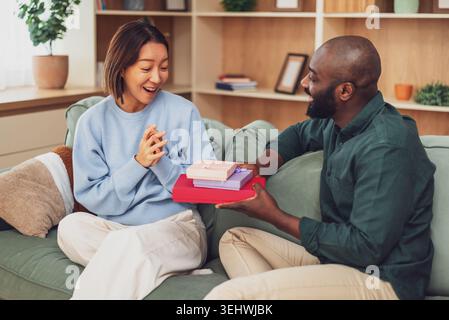 Cheerful multiracial couple is having fun at home while while exchanging gifts. Stock Photo