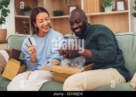 Happy multiracial couple is sitting on couch at home and shopping online. Man is holding smartphone and wife is holding payment card. Stock Photo