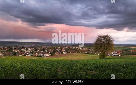 Village and rural landscape with meadows, a tree and illuminated red rain clouds at sunset Stock Photo