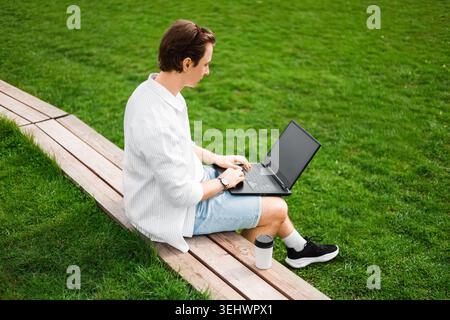 Freelancer doing remote work on a laptop outdoors. Man sitting on a wooden bench next to green grass, typing online with a coffee cup nearby. High qua Stock Photo