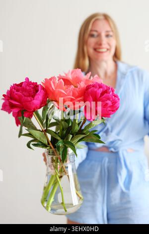Smiling young woman holding bright peonies in glass vase, soft feminine lifestyle portrait with spring flowers and natural light on minimal background Stock Photo