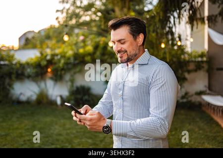 Smiling man networking using smartphone in backyard garden Stock Photo