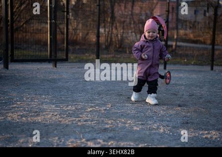 toddler in a pink beanie and purple snowsuit walking on gravel near a black metal fence at dusk, clutching small stones beside a parked stroller, warm Stock Photo