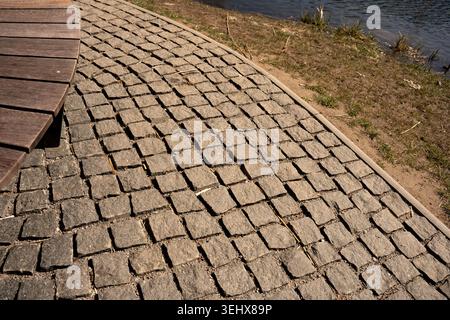 close-up view of a weathered square cobblestone pathway curving beside a wooden boardwalk and grassy riverbank with water, warm sunlight casting shado Stock Photo