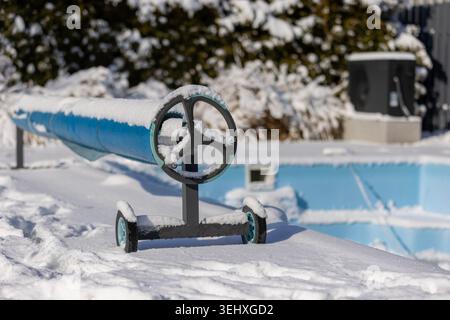 Snow-covered pool cover roller with a frozen outdoor swimming pool in the background, surrounded by a winter landscape. Stock Photo