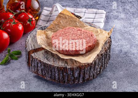 A fresh, raw ground meat burger patty on brown parchment paper, displayed on a board. Bright red tomatoes and basil add color to the gray background. Stock Photo