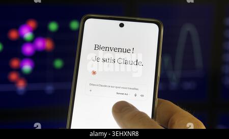 Close-up of a person's hand holding a smartphone showing the Anthropic Claude AI chatbot welcome screen in French, with a blurred compu Stock Photo