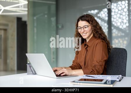 Young professional woman with glasses smiling and typing on a laptop at her desk in a contemporary office, focusing on her work and demonstrating productivity Stock Photo