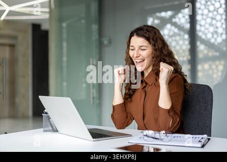 Female professional celebrating a win at her desk, fists clenched and smiling while viewing good news on her laptop in a modern office, joyful and triumphant Stock Photo