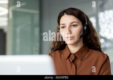 Woman in brown shirt wearing a headset, intently working at a computer screen in an office, providing professional customer support and technical assistance via video phone Stock Photo