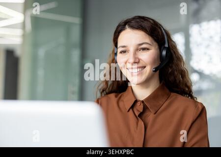 Young female call center agent wearing a headset and smiling, happy to help customers while working in a modern office environment or teleworking from home Stock Photo