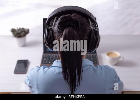 Rear view of woman wearing headphones while working at laptop desk, symbolizing online education, remote support and virtual collaboration environment Stock Photo