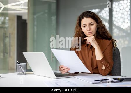 Young businesswoman with curly hair analyzing financial documents and focusing intently while working on her laptop at a desk in a modern office environment Stock Photo