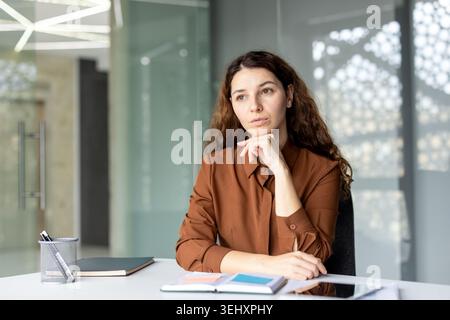 Young businesswoman with thoughtful expression and hand on chin considering new ideas and strategies while sitting at her desk in a modern corporate office environment Stock Photo