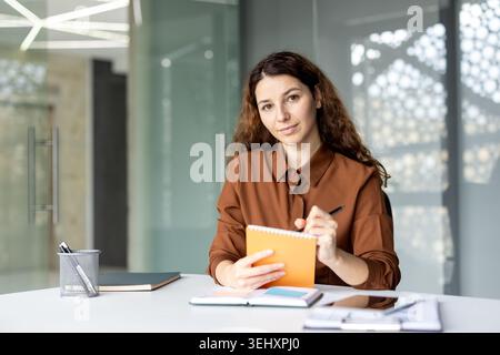 Young businesswoman with curly hair writing important notes in an orange spiral notebook, sitting attentively at a white desk in a modern office, focusing on daily tasks and professional planning Stock Photo