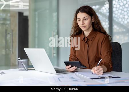 Professional woman concentrating on work, calculating financial data using a calculator and laptop, while reviewing paperwork and writing notes in a modern office setting Stock Photo