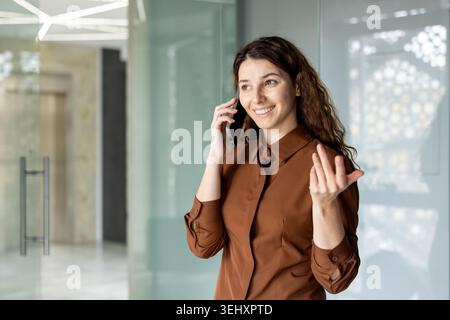 Young smiling businesswoman communicating on a mobile phone, gesturing confidently while having a professional conversation in a modern office environment Stock Photo