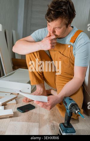 Man kneeling on the floor, intently studying paper instructions for assembling flat-pack furniture, with a cordless screwdriver, smartphone, and unassembled drawer pieces scattered around him Stock Photo