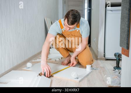 Man installing new furniture, measuring parts before assembly, completing a home improvement task, building a chest of drawers from flat pack elements on the floor Stock Photo