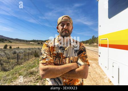 Man with a beard wearing a hawaiian shirt and cap standing with arms crossed in front of an rv on a dirt road, traveling through a rural landscape und Stock Photo