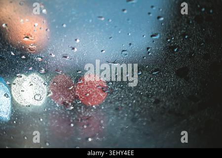 Raindrops on a window pane create a textured surface, distorting colorful street lights and city activity into soft, out of focus background bokeh, ca Stock Photo