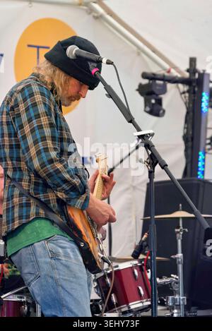 Lorton, Cumbria, United Kingdom. 18th August, 2024. A man with long blonde hair and a beard, wearing a black beanie, a plaid shirt, and jeans, plays a Stock Photo