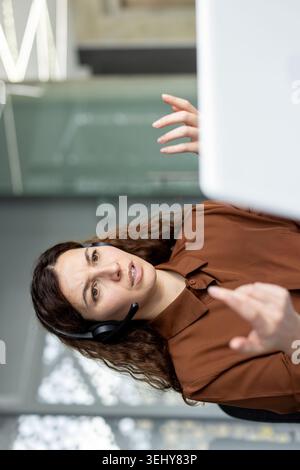Serious Latina female operator with a headset talking and gesturing during an online video call conference on a laptop in a modern corporate office room. Stock Photo