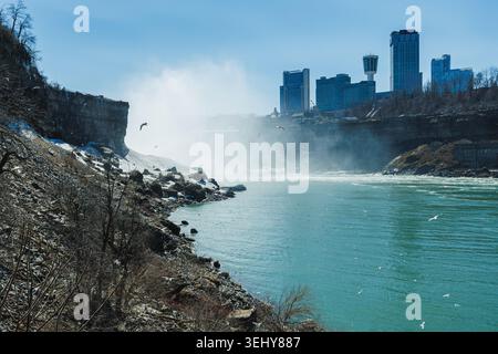 Water cascades over the edge of Niagara Falls while mist rises into the air. The citys skyline is visible nearby. Birds can be seen flying around. Stock Photo