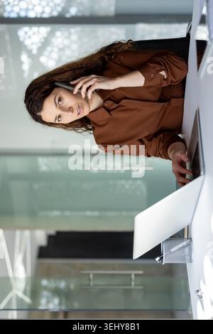 Focused Latina female manager sitting at a desk, carefully reading financial reports, paperwork documentation and analyzing data with a laptop in office. Stock Photo