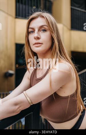 Woman leans on railing outside modern building looking directly at camera. Empowered women concept, self assured expression, authentic portraiture. Stock Photo