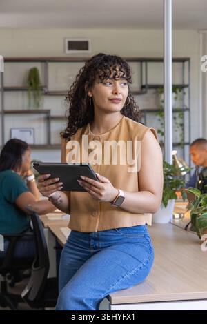 Diverse coworkers with Hispanic woman sitting on desk edge holding tablet in open-plan office Stock Photo