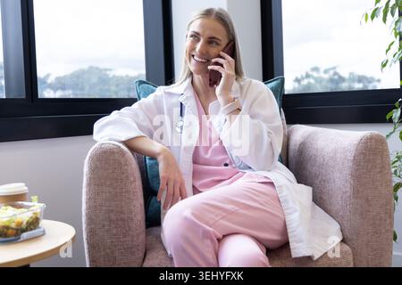 Adult female doctor sitting in clinic lounge talking on phone, wearing pink scrubs and stethoscope Stock Photo