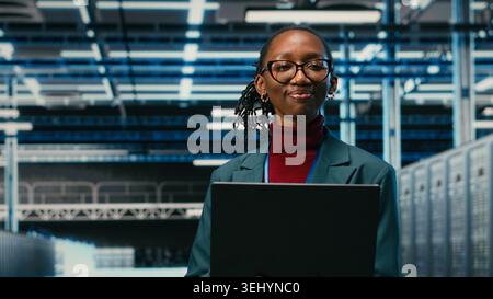 Portrait of happy data center system administrator doing resource optimization using laptop software. Smiling young employee in server hub using notebook to upgrade IT infrastructure, camera B Stock Photo
