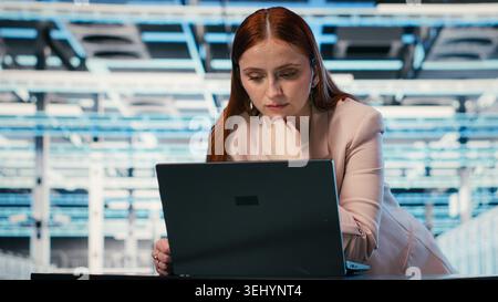 Female programmer at server room workplace desk using laptop to enhance operational effectiveness of equipment. System administrator in data center using notebook to monitor rackmounts, camera B Stock Photo