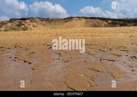 Close-up of wet sand creating geometric patterns with small pebbles on the shore. In the background, the sand dunes of Capbreton rise under a blue sky Stock Photo