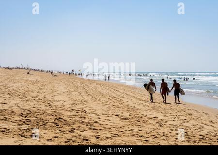 Three surfers walk along the shoreline with their boards, heading towards the ocean waves. The vast sandy beach of Capbreton is filled with many beach Stock Photo