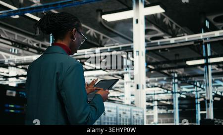 Data center programmer writing code for resource optimization using tablet application. African american woman walking in server hub using programming script on device to upgrade equipment, camera A Stock Photo