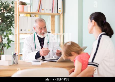 Elderly male doctor talking with mother with girl at table in medical office Stock Photo