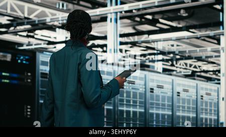 Female programmer walking in data center using tablet to write code, compiling software patches. African american woman in server hub using device application to run programming script, camera B Stock Photo